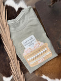 A folded sage green t-shirt with a decorative graphic of a floral pocket holding a card that reads 'God is still loving you every day.' The shirt is styled on a brown and white cowhide rug with dried palm leaves and a dark wooden surface.