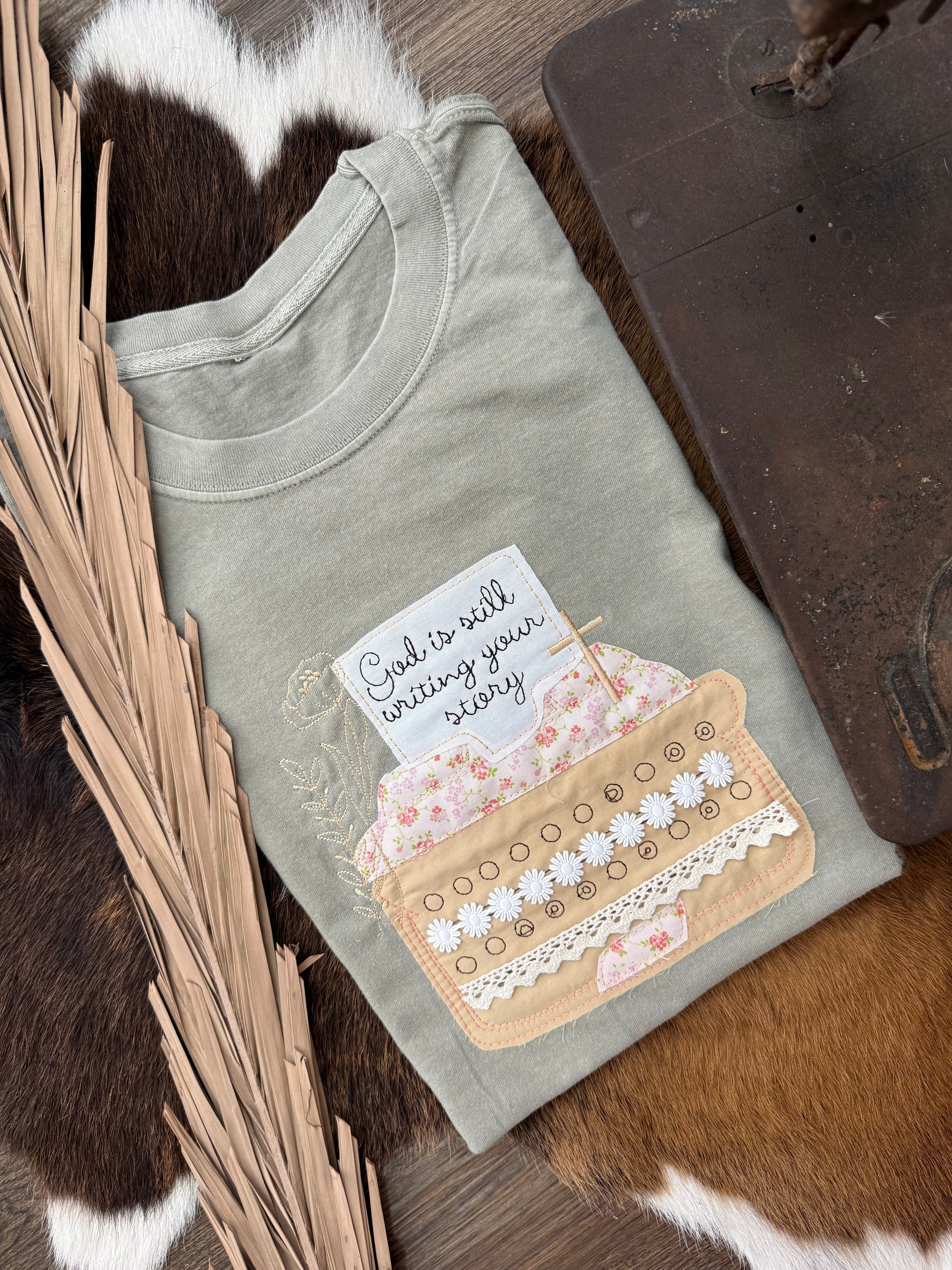 A folded sage green t-shirt with a decorative graphic of a floral pocket holding a card that reads 'God is still loving you every day.' The shirt is styled on a brown and white cowhide rug with dried palm leaves and a dark wooden surface.